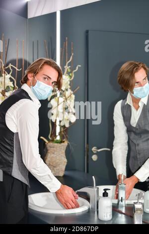Young male wearing a surgical mask and a receptionist uniform looking at the camera while washing his hands. Hygiene and safety concept. Stock Photo