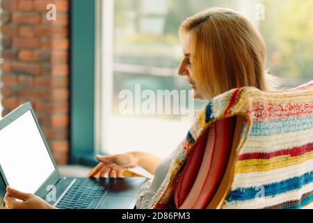 Happy woman smiling and videoconferencing on laptop while sitting in comfortable rocking chair with blurred background Stock Photo