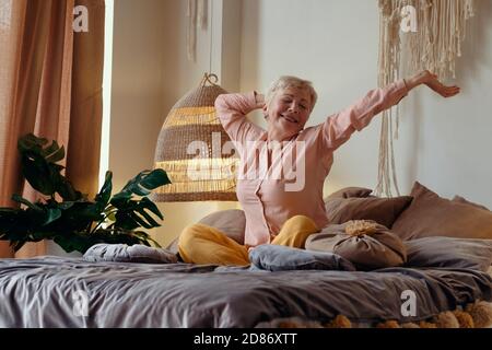 senior woman wearing pyjama smiling in bright living room stretching waking up. Stock Photo