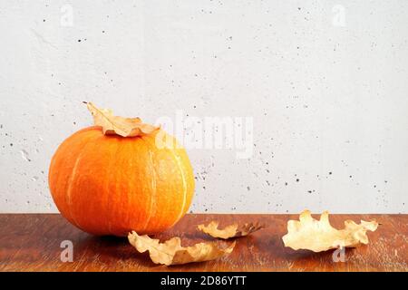 Orange pumpkin and dry oak leaves on brown wooden table on gray concrete wall background. Copy space Stock Photo