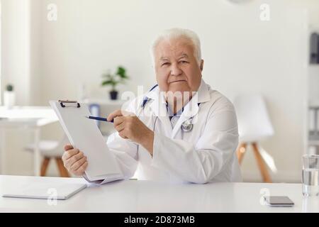 Experienced senior doctor pointing at clipboard in his hand sitting at desk in hospital office Stock Photo
