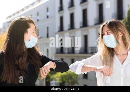 Close up of two girls greeting each other with their elbows. They are on the street and are wearing surgical masks. Concept of social distancing and n Stock Photo