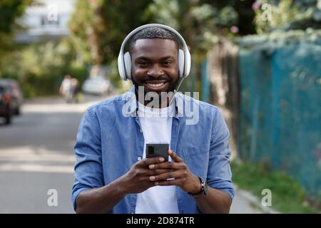Walking in the street, relaxation, leisure. Young Afro-American man in headphones listening music on smart phone using music app. Portrait of smiling Stock Photo