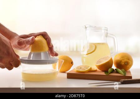 Hands squeezing a lemon in a plastic juicer with container for preparing a lemonade on the kitchen bench. Front view.Horizontal composition. Stock Photo