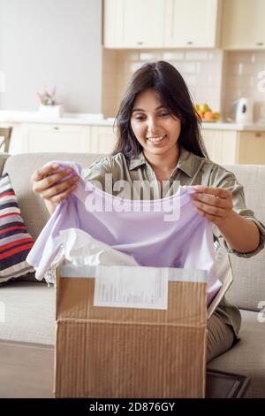 Happy indian young woman consumer opening clothes parcel box at home. Stock Photo