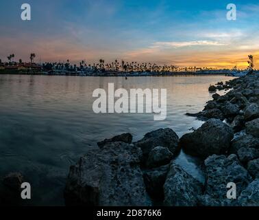 Large boulder rocks of the beach jetty creates the shoreline for the marina harbor of boats with sunrise brightening the morning sky. Stock Photo