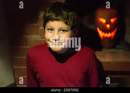 a young boy in red clothes near a brick fireplace with a Halloween jack-o'-lantern carved pumpkin with candle Stock Photo