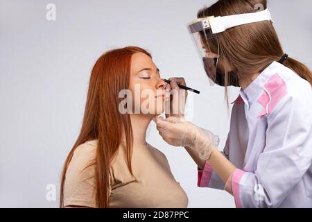 Makeup for red hair by a master dressed in medical robe, mask, gloves, protective screen . New normal beauty concept.  Stock Photo