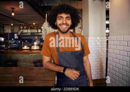 Portrait of smiling young male waiter wearing apron leaning on table standing looking at camera in cafe Stock Photo