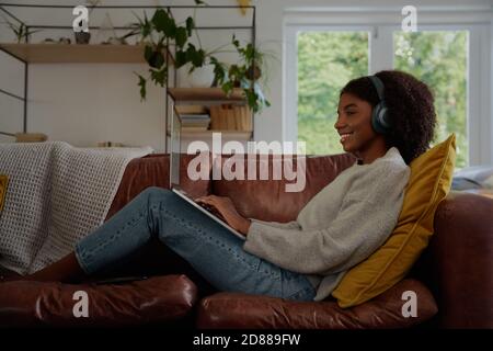 Attractive young woman using a laptop while listening to music on headphones and relaxing on a sofa at home Stock Photo