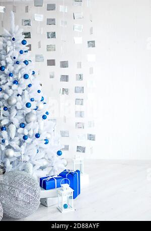 Christmas white artificial fir tree in a light gray room is decorated with blue and silver balloons. Boxes with gifts, decorations made of cubes on th Stock Photo