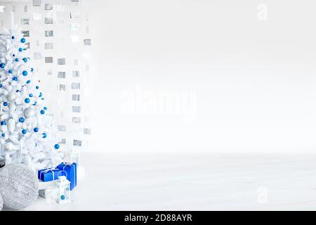 Christmas white artificial fir tree in a light gray room is decorated with blue and silver balloons. Boxes with gifts, decorations made of cubes on th Stock Photo