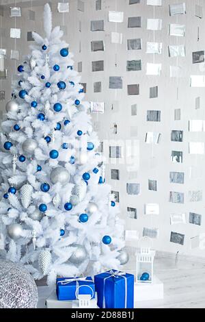 Christmas white artificial fir tree in a light gray room is decorated with blue and silver balloons. Boxes with gifts, decorations made of cubes on th Stock Photo