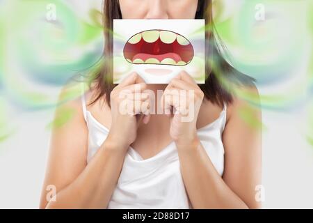 A woman wearing white dress holding a white paper with an open mouth cartoon image. On a light gray background. Bad breath or Halitosis. The concept w Stock Photo