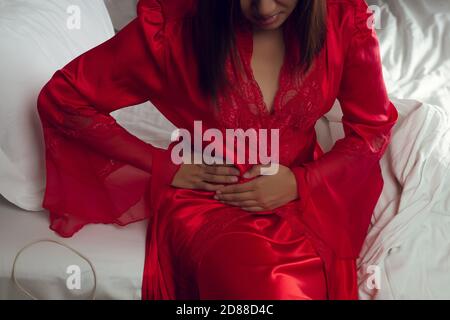 A woman in satin red nightgown suffering from menstruation pain on the bed at night. Irritable Bowel Syndrome or IBS Stock Photo