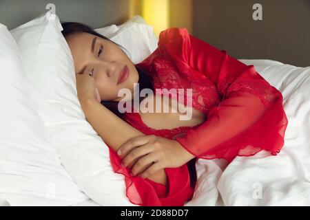 Tired woman lying in bed can't sleep late at night with insomnia. Girl wore a satin nightgown and red robe lying on the bed awake in the bedroom Stock Photo
