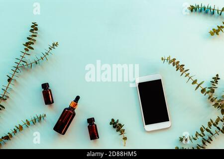 Essential oils and serum in dark glass bottles next to the mobile phone, eucalyptus branches on light blue background. Natural cosmetics and aromather Stock Photo