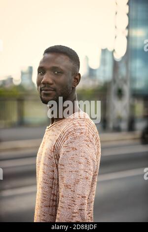 Portrait of handsome black man turning head looking at camera. City background. Shallow depth of field. Medium shot. Stock Photo