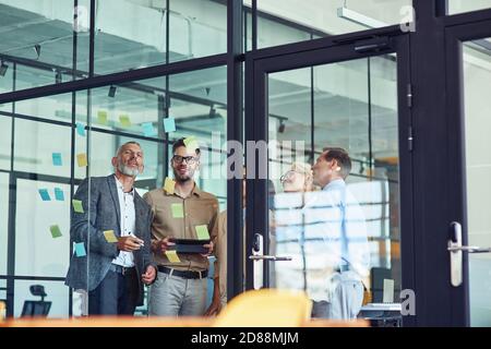 Group of creative coworkers standing in the modern office and looking at colorful sticky notes on a glass wall, discussing project Stock Photo