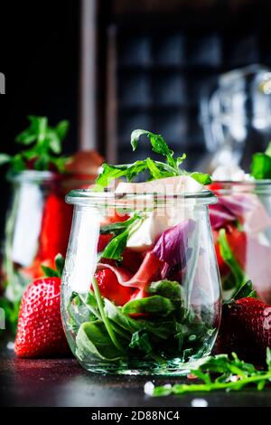 Summer salad with arugula, soft cheese, red strawberry and prosciutto in glass jars on black table, selective focus Stock Photo