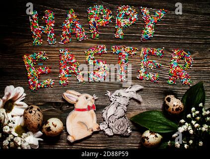 Vintage Easter wooden background with the text "Happy Easter", made of multi-colored sweet topping for Easter cakes and painted cardboard rabbits, top Stock Photo
