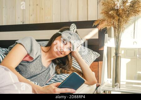 Woman in sleep mask make morning selfie in bed. Striped gray bedding, wooden wall. Sun light on the wall. Stock Photo