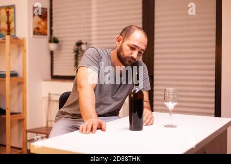 Young man being lonely in home having alcohol addiction sitting at table in kichen. Unhappy person disease and anxiety feeling exhausted with having alcoholism problems. Stock Photo