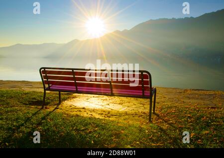 Bench with Sunbeam on Misty Alpine Lake Maggiore with Mountain in Ascona, Ticino in Switzerland. Stock Photo