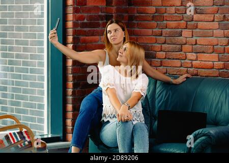Relaxed blonde girlfriends are sitting on the sofa in the living room. One of the women takes a selfie. Big city life Stock Photo