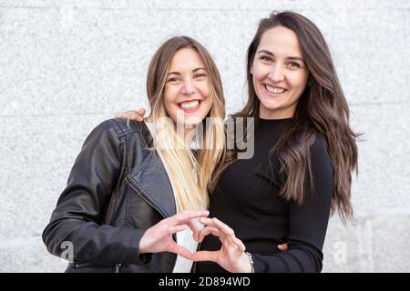 Two smiling girls hugging and forming a heart with their hands. They are out in the open. Stock Photo