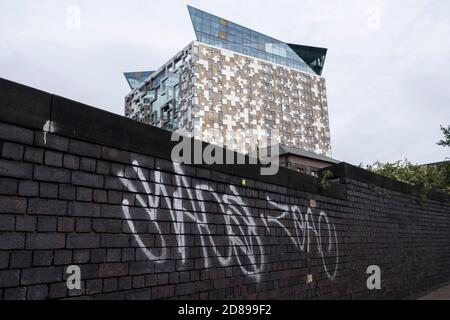 Exterior detail of The Cube building behind an old wall covered in graffiti on 7th October 2020  in Birmingham, United Kingdom.  The Cube is a 25 storey mixed-use development in the centre of Birmingham, England. Designed by Ken Shuttleworth of MAKE Architects, it contains 135 flats, offices, shops, a hotel and a restaurant. It is the final phase of The Mailbox development. Stock Photo