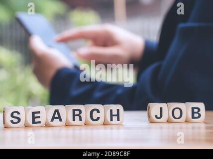 Closeup wooden block written Jobs Search on table against bussinessman using smart phone to find new jobs and application. Employment concept. Stock Photo