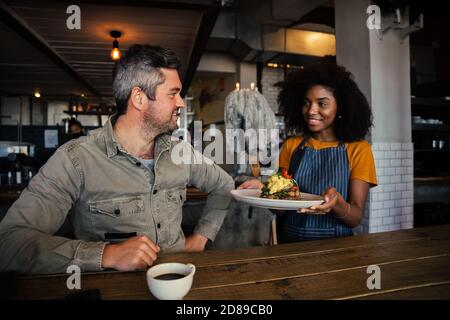 Smiling waitress serving male customer fresh egg breakfast while he drinks hot coffee sitting in funky cafe  Stock Photo