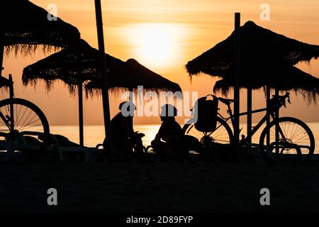 Silhouette of two cyclists sitting on beach under parasols at sunrise, Fuengirola, Malaga, Spain Stock Photo