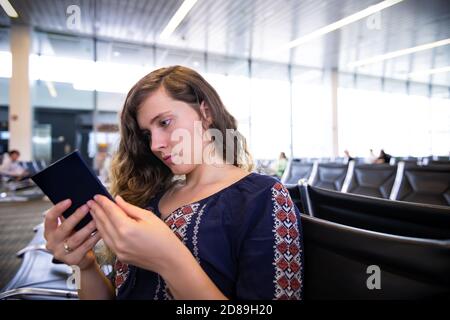 Closeup portrait of young woman girl holding looking at blue American United States of America passport sitting in airport terminal gate waiting for a Stock Photo