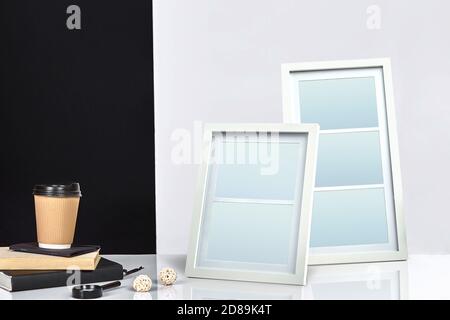 Two blank frames, paper cup of coffee on books and magnifier on tabletop against black and white studio background. Close up, copy space Stock Photo