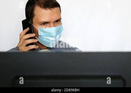 Man in face mask and office clothes talking on mobile phone in front of PC monitor. Safety at work during coronavirus pandemic Stock Photo