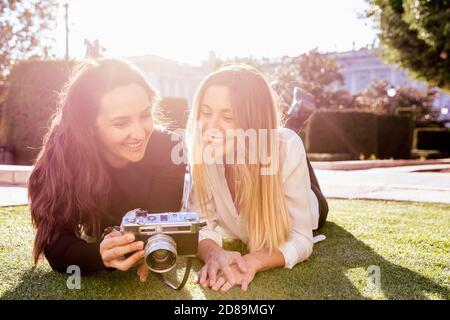 Two women lying on the grass smile as they look at a vintage camera. Ellas tienen el sol a sus espaldas. Stock Photo