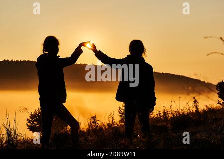 Beautiful fog in the morning. Silhouettes of two girls making heart with their hands against a beautiful golden sunrise. Stock Photo