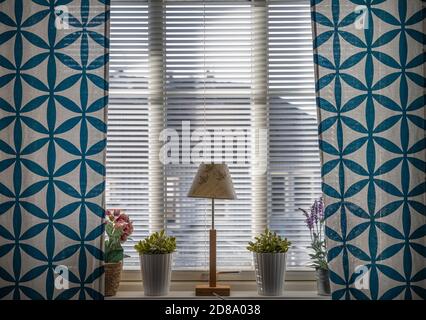 A window with blue curtains, a lamp and flowers. Beautiful light comes in through the blinds Stock Photo