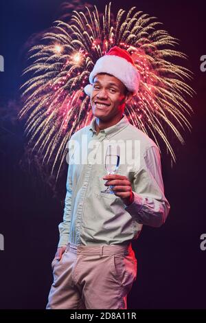 Happy New Year. Vertical shot of a young cheerful mixed race man wearing Santa Claus cap with glass of champagne standing against fireworks background and smiling. Christmas party, people and holidays Stock Photo