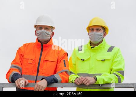 Two workers or engineers with face mask standing on a construction site or  an industrial railing - bright clear background Stock Photo
