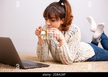 Portrait of young woman lying on floor with laptop computer and drinking tea Stock Photo