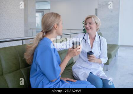 Two smiling female medical workers sitting on sofa, drinking coffee Stock Photo