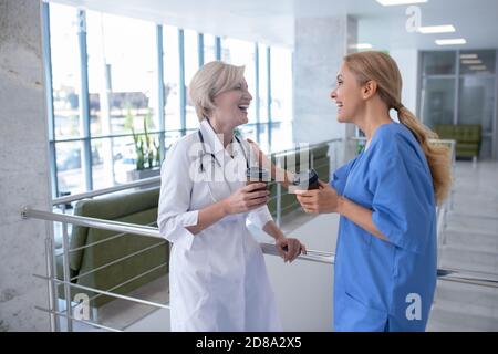 Two smiling female medical workers leaning on stair railing, drinking coffee Stock Photo