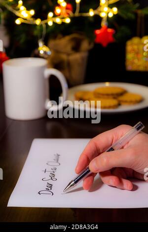 Close-up of a woman's hand writing a letter for Santa Claus. In the background cup of milk, a plate with cookies and a Christmas tree. Christmas and h Stock Photo