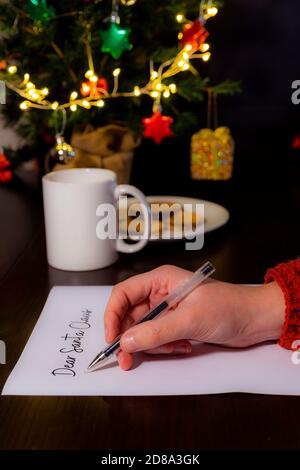Close-up of a woman's hand writing a letter for Santa Claus. In the background cup of milk, a plate with cookies and a Christmas tree. Christmas and h Stock Photo