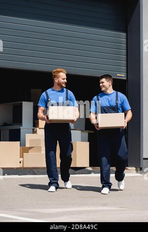 Movers in overalls looking at each other while holding cardboard boxes on urban street Stock Photo