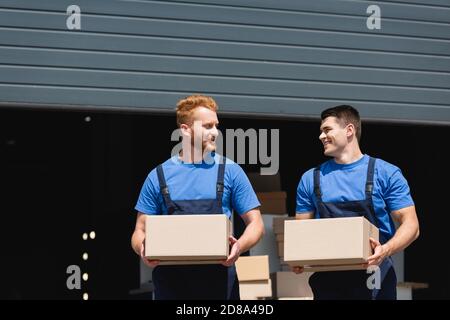 Young movers in uniform looking at each other while holding cardboard boxes on urban street Stock Photo