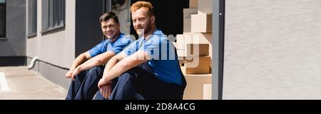 Panoramic shot of movers looking at camera while sitting in warehouse outdoors Stock Photo